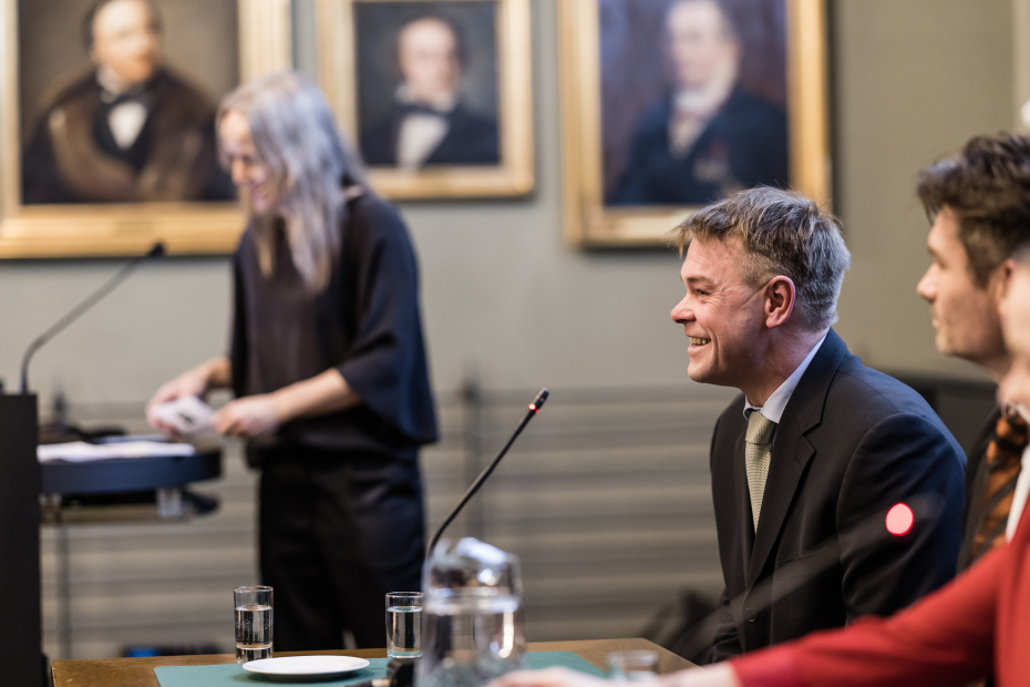 Smilende mann i profil med dame utenfor fokus i bakgrunnen.