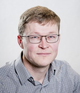 Blond mann i grå skjorte smiler forsiktig mot kamera. Fotografiet er tatt i et studio med hvit bakgrunn.
