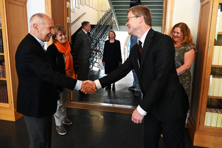 Two men in dark suits meeting each other with a handshake. They are smiling.